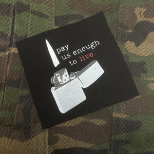 May include: A black square patch with the text "I pay us enough to live." and a white illustration of a lighter. The patch is set against a camouflage background.