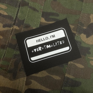 Puede incluir: Primer plano de una chaqueta de camuflaje con un parche rectangular negro. El parche muestra el texto blanco "HELLO, I'M OVERSTIMULATED" dentro de un borde blanco. El patrón de la chaqueta incluye tonos verdes, marrones y negros.