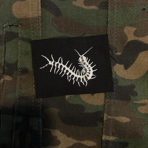 May include: A camouflage-patterned fabric with a black rectangular patch. The patch features a white, stylized centipede design. The fabric has a mix of green, brown, and black colors.