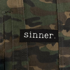 Könnte beinhalten: Nahaufnahme einer Camouflage-Jacke mit einem schwarzen Aufnäher. Der Aufnäher zeigt das Wort "sinner." in weißer, serifenloser Schrift. Das Muster der Jacke umfasst Grün-, Braun- und Schwarztöne.