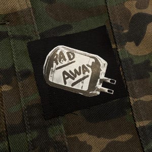 May include: Camouflage jacket with a black patch. The patch features the words "RAD AWAY" in white, stylized text, above a white graphic. The jacket has a green, brown, and black pattern.