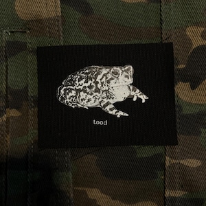 May include: A black patch with a white illustration of a toad and the word "tood" beneath it. The patch is set against a camouflage pattern background in shades of green and brown.