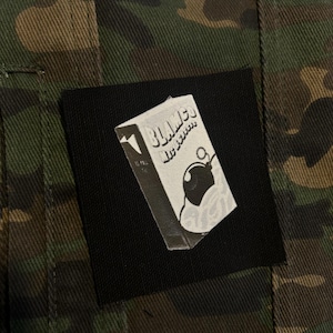 May include: A black square patch featuring a grayscale illustration of a carton with the word "Blame" and a bomb design. The patch is set against a camouflage pattern background.