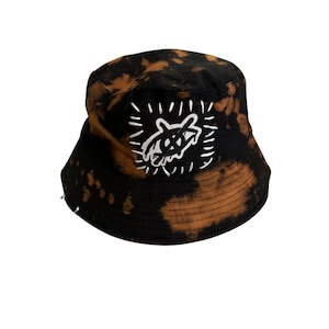 Peut inclure: Bob noir avec un effet tie-dye marron et noir. Un patch carré blanc est cousu sur le devant, avec un motif graphique blanc. Le chapeau a une forme de chapeau de pêcheur classique.