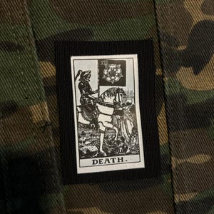May include: A rectangular fabric patch featuring a black and white illustration of the Death tarot card, depicting a knight on a horse holding a flag with a rose emblem, sewn onto a green, brown, and black camouflage patterned textile background.