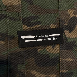 Puede incluir: Una tela con patrón de camuflaje que presenta una etiqueta rectangular negra cosida en la superficie. La etiqueta muestra el texto blanco "trust all authority" con gráficos de pinceladas blancas horizontales encima y debajo del texto.