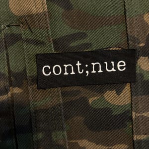 May include: A fabric garment featuring a green, brown, and black camouflage pattern. A rectangular black fabric label is stitched onto the material, displaying the white lowercase text "cont;nue" in a serif font, positioned horizontally.