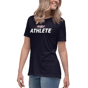 May include: A dark navy blue short-sleeved t-shirt featuring the word ATHLETE in large white capital letters. Above the word ATHLETE, the word FEMALE is printed in a smaller font with a red line struck through it.