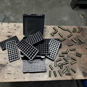 Puede incluir: Un conjunto de bandejas de almacenamiento de munición de plástico negro y casquillos de bala de latón sobre una superficie de madera. Las bandejas, con múltiples agujeros, están abiertas y cerradas. Los casquillos son cilíndricos.