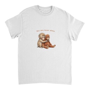 May include: White t-shirt featuring a heartwarming illustration of two primates embracing. The text "you are never alone" is printed above the image. The design is centered on the front of the shirt.