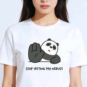 May include: White t-shirt with a cartoon panda illustration. The panda has a grumpy expression and is raising a hand. The text "STOP HITTING MY NERVES" is printed below the panda. The shirt is made of a soft material.