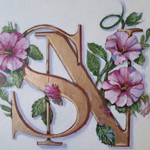 Puede incluir: Un monograma dorado con las letras "SN" entrelazadas, adornado con flores de hibisco rosa y hojas verdes. La obra de arte está sobre un fondo blanco, creando un contraste que resalta los detalles florales y de las letras.