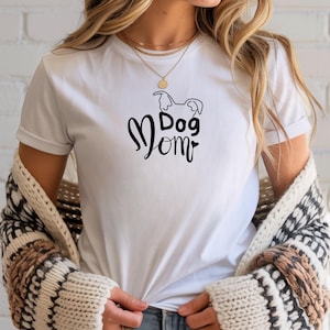 Könnte beinhalten: Weißes T-Shirt mit dem Schriftzug "Dog Mom" in schwarzer Schrift, ergänzt durch eine Hunde-Silhouette. Das Shirt hat kurze Ärmel und einen Rundhalsausschnitt.