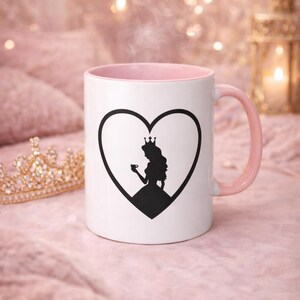 Könnte beinhalten: Weiße Keramiktasse mit rosa Innenraum und Griff. Eine schwarze Herzform auf der Vorderseite enthält eine Silhouette einer Prinzessin, die eine Teetasse hält. Eine goldene Krone befindet sich links von der Tasse. Dampf steigt aus der Tasse auf.