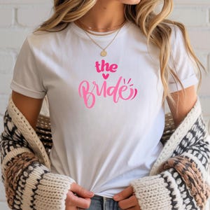 Könnte beinhalten: Weißes T-Shirt mit dem Text "the Bride" in rosa Schrift. Das Wort "the" ist fett und ohne Serifen, mit einem rosa Herz über dem Wort "Bride". Das Shirt hat kurze Ärmel.