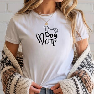 Könnte beinhalten: Weißes T-Shirt mit dem Schriftzug "Dog Mom" in schwarzer Schrift, ergänzt durch eine Hunde-Silhouette. Das Shirt hat kurze Ärmel und einen Rundhalsausschnitt.