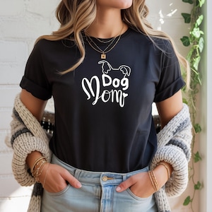 Könnte beinhalten: Schwarzes T-Shirt mit den Worten "Dog Mom" in weißer Schrift und einer Hundeumrisslinie. Das Shirt hat hochgekrempelte Ärmel und wird mit Jeans kombiniert. Eine Frau trägt das Shirt mit einer mehrschichtigen Halskette und Armbändern.