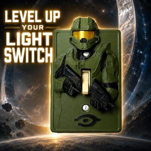 May include: A rectangular olive green light switch cover featuring a molded relief of a soldier in armor with a gold visor. The center includes a cream-colored toggle switch and a screw. The text LEVEL UP YOUR LIGHT SWITCH is printed on the left.