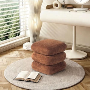 May include: Three stacked, square, brown cushions on a light gray, round rug. An open book rests on the rug near the cushions. The scene is set in a room with a white desk, a decorative lamp, and a window with blinds.