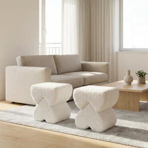 Peut inclure: Deux poufs crème texturés en forme de sablier sont posés sur un tapis gris devant un canapé beige. Une table basse en bois clair et un vase complètent le décor d'un salon moderne.