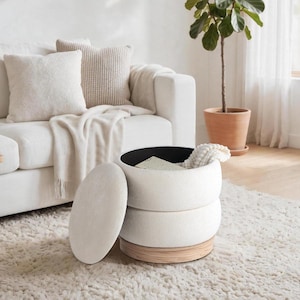 May include: A white, round storage ottoman with a light wood base and a black interior. The lid is off, revealing the inside. The ottoman is next to a white sofa with cushions and a throw. A potted plant is in the background.