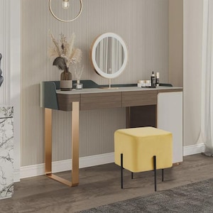 May include: A modern vanity set with a wooden desk, gold legs, and a round lighted mirror. The desk has two drawers and a white cabinet. A yellow velvet stool with black legs sits in front of the desk. Decorative items are on the desk.