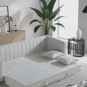 Op de afbeelding: Een lichtgrijze slaapbank met twee bijpassende vierkante kussens. De bank staat in een moderne woonkamer met een zwart bijzettafeltje, een vloerlamp en een grote potplant. De bank is uitgetrokken tot bed.
