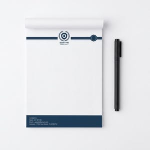 May include: A white notepad with a dark blue header featuring a shield logo and the text Security Pro Consultancy, and a dark blue footer with contact details including a phone number, email, and address. A black pen is positioned to the right.