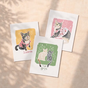 May include: Three rectangular art prints featuring cat illustrations. One print shows a cat in a pink polka dot shirt labelled Isabelle. Another shows a cat in a floral collar on a pink background. The third shows two cats on a green background labelled The Boys.