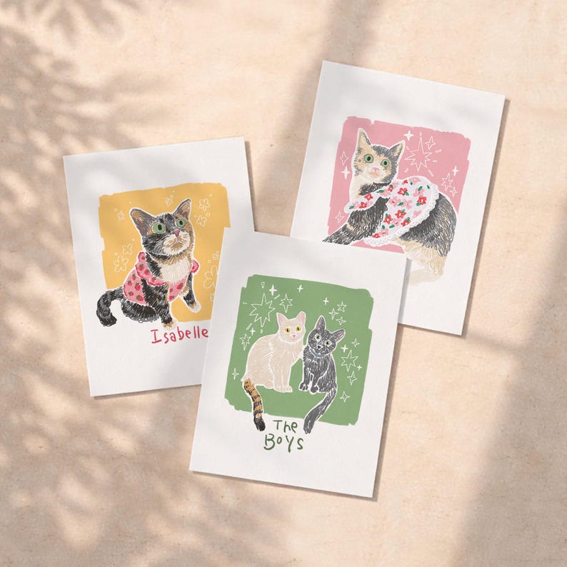 May include: Three rectangular art prints featuring cat illustrations. One print shows a cat in a pink polka dot shirt labelled Isabelle. Another shows a cat in a floral collar on a pink background. The third shows two cats on a green background labelled The Boys.