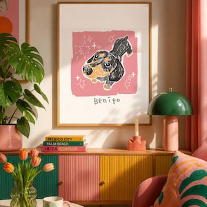 May include: A framed wall art print featuring a black and tan dachshund illustration on a pink background with white star accents and the name Benito written in black ink below the dog. The artwork is displayed in a simple wooden frame.