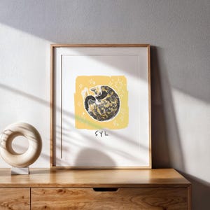 May include: A framed art print featuring a curled-up tortoiseshell cat centred on a yellow square background with white star illustrations. The text SYL is printed in black below the cat. The artwork is enclosed in a light wood frame.