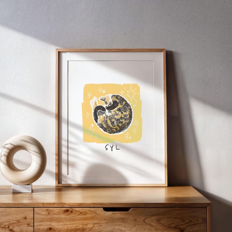 May include: A framed art print featuring a curled-up tortoiseshell cat centred on a yellow square background with white star illustrations. The text SYL is printed in black below the cat. The artwork is enclosed in a light wood frame.