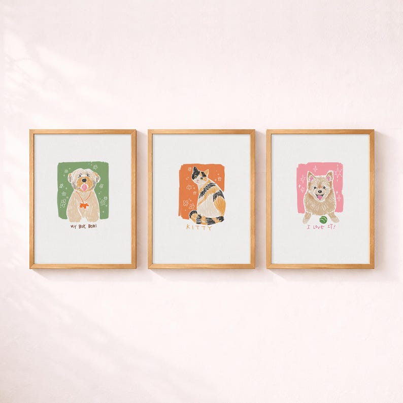 May include: Three framed wall art prints featuring illustrations of pets. The left print shows a tan dog on a green background with the text MY BOY BORI. The centre print shows a calico cat on an orange background with the text KITTY. The right print shows a tan dog with a ball on a pink background with the text I LOVE IT.