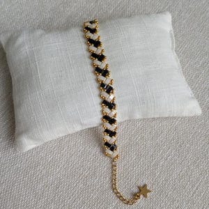 Peut inclure: Un bracelet de perles avec une chaîne dorée et un charm en forme d'étoile. Le bracelet présente un motif répété de perles noires et blanches, séparées par des perles dorées. Le bracelet est présenté sur un coussin blanc.