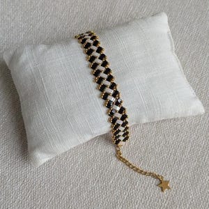 Peut inclure: Un bracelet à motifs noirs et blancs, orné de perles dorées. Le bracelet est présenté sur un coussin blanc et comporte une chaîne dorée avec un charm en forme d'étoile.