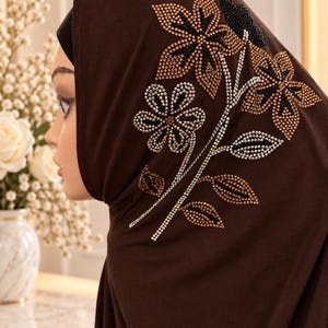 May include: A dark brown hijab featuring a decorative floral pattern composed of small, shimmering rhinestones in shades of gold, silver, and black. The design includes three distinct flower heads and several leaves arranged on a single stem.
