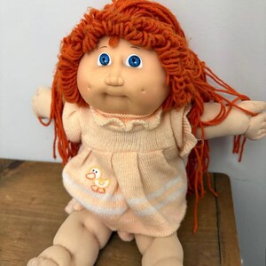 Könnte beinhalten: Vintage Cabbage Patch Kid Puppe mit orangefarbenem Garnhaar und blauen Augen. Die Puppe trägt ein pfirsichfarbenes Kleid mit einem Entenmuster und weißen Streifen. Die Puppe sitzt auf einer Holzoberfläche.