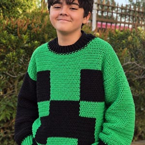 Könnte beinhalten: Ein langärmeliger Häkelpullover mit einem pixeligen grün-schwarzen Blockmuster auf der Vorderseite, einem einfarbigen schwarzen gerippten Rundhalsausschnitt, schwarzen gerippten Bündchen und einem schwarzen gerippten Saum, mit einem schwarzen und einem grünen Ärmel.