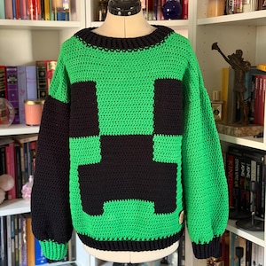Könnte beinhalten: Ein langärmeliger Häkelpullover mit einer leuchtend grünen Basis und einem großen, schwarzen, verpixelten Gesichtsmuster auf der Vorderseite. Das Kleidungsstück hat einen dicken schwarzen Rippstrickkragen, schwarze Bündchen und einen schwarzen Rippsaum.