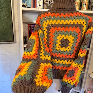 Puede incluir: Un suéter de cuello alto marrón hecho a mano con un patrón geométrico colorido. El patrón presenta cuadrados de color naranja, amarillo y verde. Las mangas son largas y abullonadas. El suéter se exhibe en un maniquí.