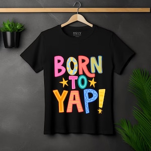 May include: Black t-shirt featuring the text "BORN TO YAP!" in vibrant, multi-coloured letters. The design incorporates stars and a playful font, creating a cheerful and engaging graphic tee.