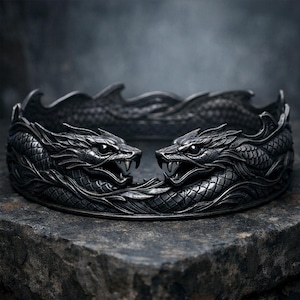 May include: A dark grey bracelet with two dragon heads facing each other. The dragons have open mouths with visible teeth and are surrounded by intricate scale and wave patterns. The bracelet is made of a dark metal and sits on a textured surface.