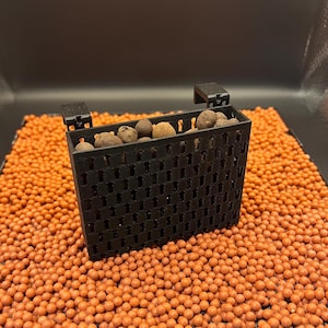 May include: A rectangular black plastic planter basket with a grid-like perforated design and two top hooks, filled with small brown clay pebbles. The planter sits on a flat surface covered in a layer of numerous small, round, orange clay pellets.