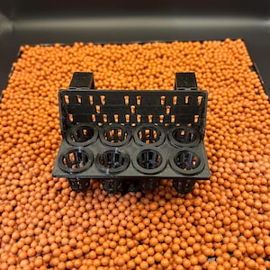 Op de afbeelding: Een zwarte plastic hydroponische plantenhouder met acht cirkelvormige openingen, ontworpen om kleine planten vast te houden. De houder heeft een rechthoekige vorm met een rasterpatroon en staat tegen een achtergrond van oranje kleikorrels.