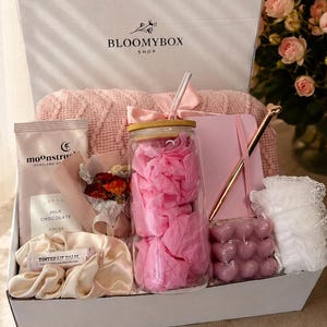 May include: A white gift box labelled BLOOMYBOX SHOP containing a pink knit blanket, a glass tumbler with a bamboo lid and straw, a pink notebook with a gold pen, a chocolate bar, a tinted lip balm, a silk scrunchie, and a heart-shaped candle.