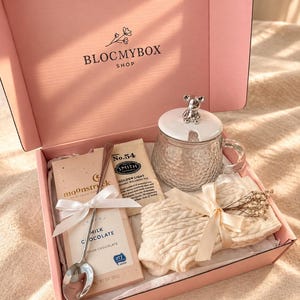May include: A pink gift box labelled BLOCMYBOX SHOP containing a glass mug with a bear-topped lid, a bar of Moonstruck milk chocolate, a Smith No. 54 tea packet, a silver spoon, and a cream-coloured cable-knit item tied with a ribbon.