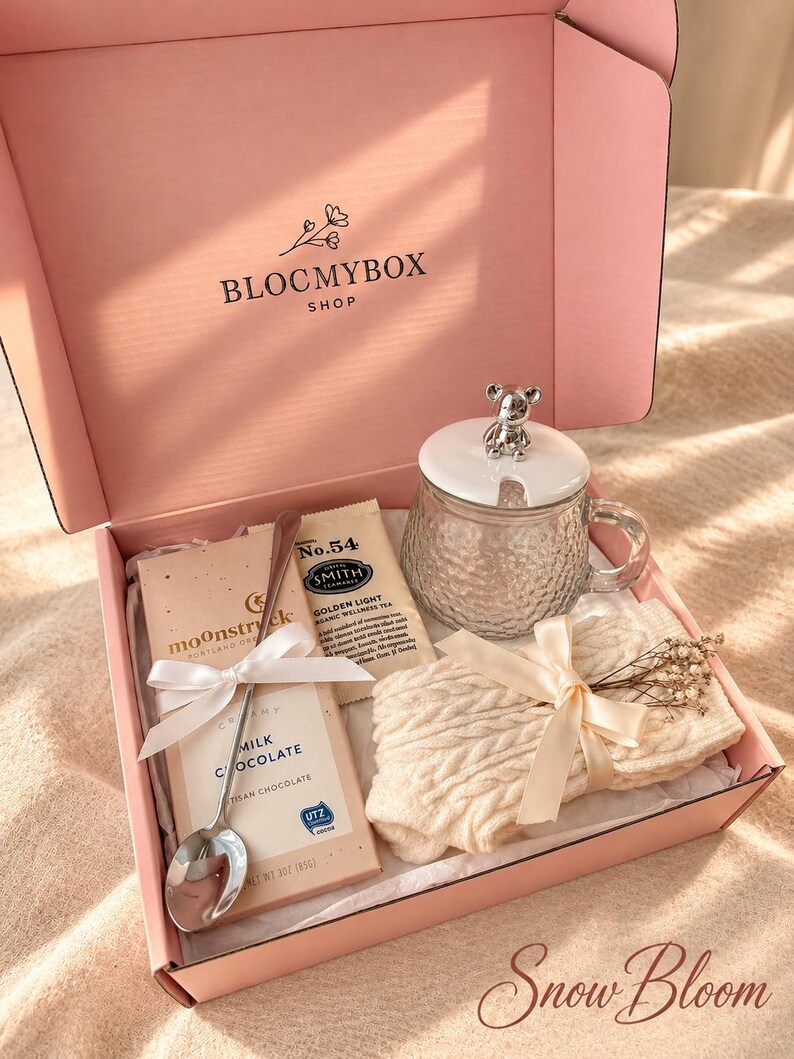 May include: A pink gift box labelled BLOCMYBOX SHOP containing a glass mug with a bear-topped lid, a bar of Moonstruck milk chocolate, a Smith No. 54 tea packet, a silver spoon, and a cream-coloured cable-knit item tied with a ribbon.