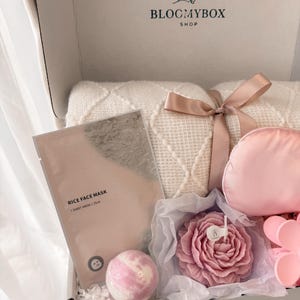 Puede incluir: Caja de regalo abierta con una manta blanca atada con una cinta, una mascarilla facial rosa, una bomba de ba&ntilde;o, una vela con forma de rosa y una m&aacute;scara de ojos rosa. La caja tiene el texto "BLOCMYBOX SHOP".