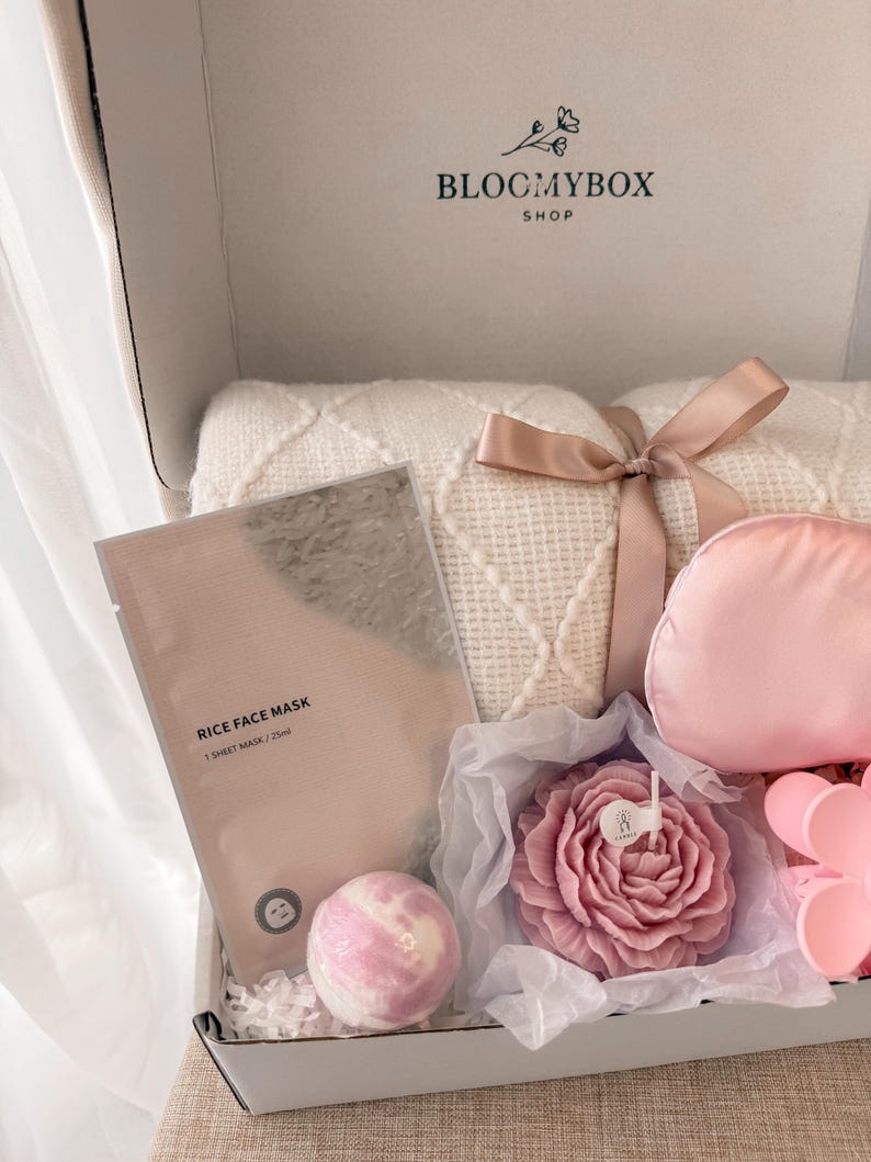 Puede incluir: Caja de regalo abierta con una manta blanca atada con una cinta, una mascarilla facial rosa, una bomba de ba&ntilde;o, una vela con forma de rosa y una m&aacute;scara de ojos rosa. La caja tiene el texto "BLOCMYBOX SHOP".